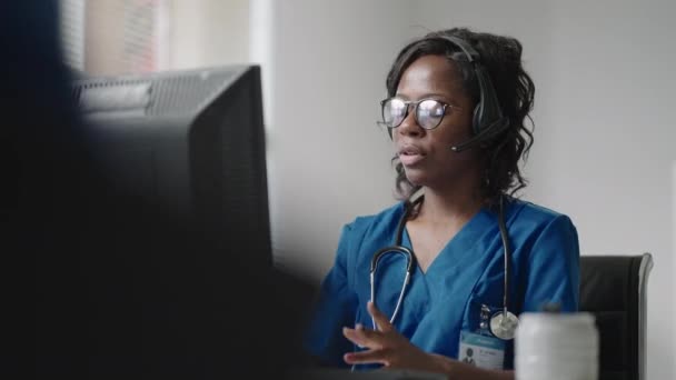 Uma médica negra usando fones de ouvido se senta em uma mesa com um computador e recebe chamadas de pacientes olha para seus registros médicos e os insere no cronograma da clínica. — Vídeo de Stock