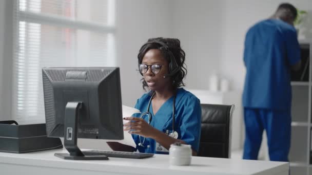 ヘッドフォンを装着した黒人女性医師がコンピュータを持ってテーブルに座り、患者からの電話で診療記録を見て診療所に入る。 — ストック動画