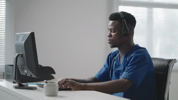 Afro-américain mâle médecin ou infirmière avec casque et ordinateur travaillant à l'hôpital. Jeune thérapeute professionnel médecin consultant client en utilisant la communication à distance parlant sur webcam — Video