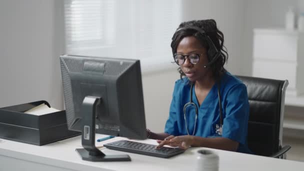 Afrikaanse vrouwelijke medisch assistent draagt witte jas, headset video bellen verre patiënt op de computer. Arts praten met de klant met behulp van virtuele chat computer app. Telegeneeskunde, gezondheidszorg op afstand — Stockvideo