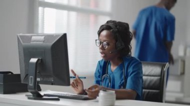 Beyaz önlüklü Afrikalı siyahi kadın pratisyen doktor muayenehanesinde masa başında oturmuş hastaların tıbbi geçmişini okurken bilgisayar faresi kaydırıyor.