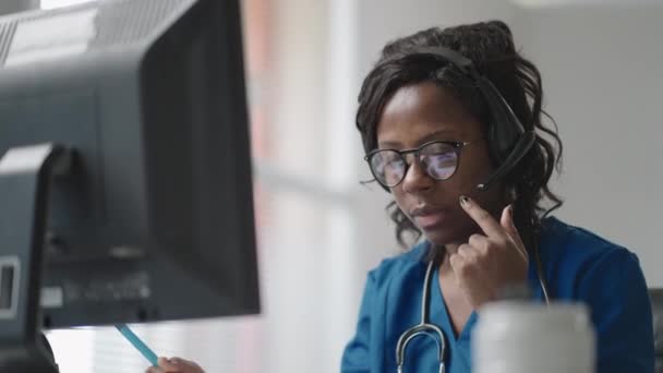 Mujer africana asistente médico lleva una bata blanca, auriculares de vídeo llamando a paciente distante en el ordenador. Doctor hablando con el cliente utilizando la aplicación de chat virtual de la computadora. Telemedicina, asistencia sanitaria a distancia — Vídeos de Stock