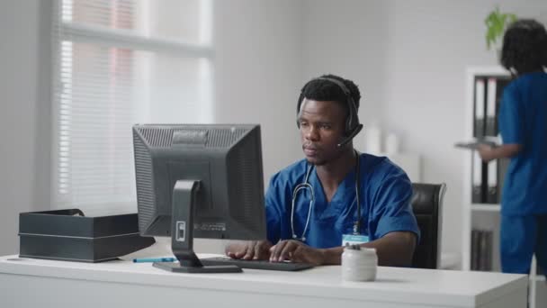 Afro-americký lékař nebo zdravotní sestra se sluchátky a počítač pracuje v nemocnici.young profesionální terapeut lékař poradenství zákazník klient pomocí dálkové komunikace mluvení na webkameru — Stock video
