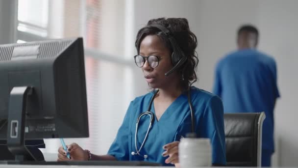 Африканська жінка-медик носить біле пальто, відео з навушниками телефонує віддаленому пацієнту на комп'ютері. Доктор розмовляє з клієнтом за допомогою віртуального комп'ютерного додатку. Телемедицина, віддалене медичне обслуговування — стокове відео
