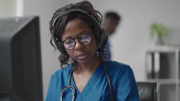 Чорношкіра жінка-лікар у навушниках сидить за столом з комп'ютером і приймає дзвінки від пацієнтів, дивлячись на їх медичні записи і входить в їхній розклад — стокове відео