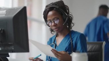 Beyaz önlüklü Afrikalı siyahi kadın pratisyen doktor muayenehanesinde masa başında oturmuş hastaların tıbbi geçmişini okurken bilgisayar faresi kaydırıyor.