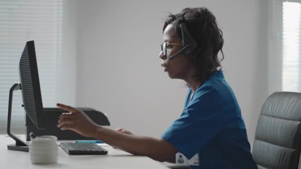 Afrikansk amerikansk svart kvinnlig allmänläkare i vit rock sitter vid skrivbordet i läkare kontor och rulla datormus när du läser patienter sjukdomshistoria — Stockvideo