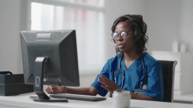 Beyaz önlüklü Afrikalı siyahi kadın pratisyen doktor muayenehanesinde masa başında oturmuş hastaların tıbbi geçmişini okurken bilgisayar faresi kaydırıyor.