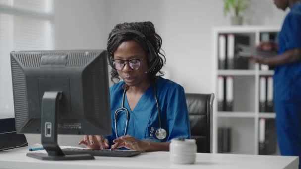 Assistente médica feminina africana usa casaco branco, fone de ouvido de vídeo chamando paciente distante no computador. Médico falando com o cliente usando aplicativo de computador de bate-papo virtual. Telemedicina, cuidados de saúde remotos — Vídeo de Stock