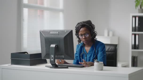 Una doctora negra que usa auriculares se sienta en una mesa con una computadora y recibe llamadas de pacientes mira sus registros médicos y los ingresa en el programa de la clínica. — Vídeo de stock