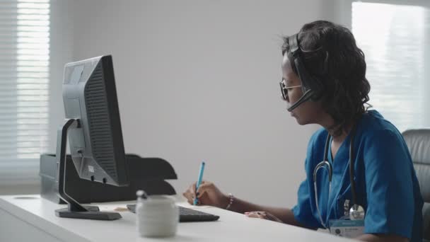 Assistente médica feminina africana usa casaco branco, fone de ouvido de vídeo chamando paciente distante no computador. Médico falando com o cliente usando aplicativo de computador de bate-papo virtual. Telemedicina, cuidados de saúde remotos — Vídeo de Stock