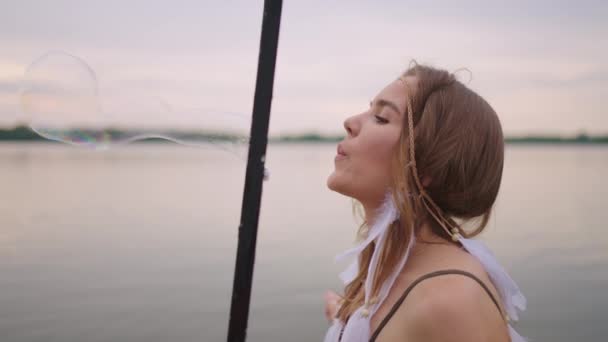 Une jeune artiste féminine souffle beaucoup de bulles de savon, montre un spectacle théâtral en utilisant un cadre au ralenti au coucher du soleil sur un lac — Video