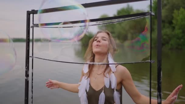 En ung kvindelig kunstner blæser en masse sæbebobler, viser et teatershow ved hjælp af en ramme i slowmotion ved solnedgang på en sø – Stock-video