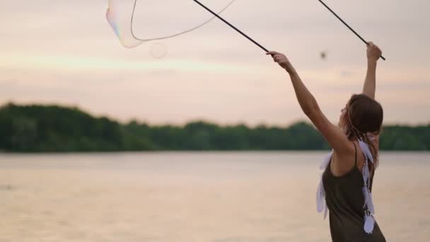 En ung hippie kvinna i klänning och med fjädrar på huvudet gör enorma såpbubblor vid solnedgången på stranden av en sjö i slow motion — Stockvideo