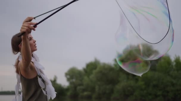 A young hippie woman in a dress and with feathers on her head makes huge soap bubbles at sunset on the shore of a lake in slow motion — Stock Video