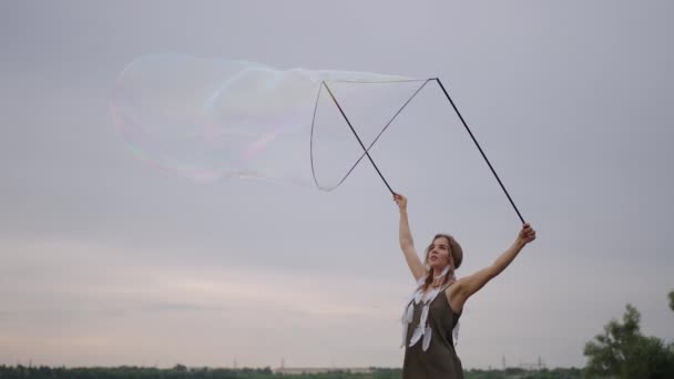 Een jonge hippie vrouw in een jurk en met veren op haar hoofd maakt enorme zeepbellen bij zonsondergang op de oever van een meer in slow motion — Stockvideo