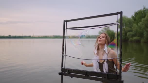 Una joven artista sopla un montón de pompas de jabón, muestra un espectáculo teatral usando un marco en cámara lenta al atardecer en un lago — Vídeo de stock