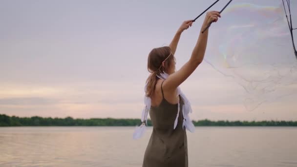 一个身穿裙子头戴羽毛的年轻嬉皮士女人在日落时分缓慢地在湖岸上制造巨大的肥皂泡 — 图库视频影像
