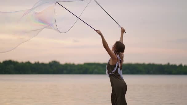 Egy fiatal női művész egy szappanbuborék show-t mutat, ami hatalmas szappanbuborékokat robbant fel egy tó partján naplementekor. Mutasd meg a gyönyörű show szappanbuborékok lassított felvételen — Stock videók