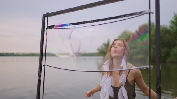 Una giovane artista femminile soffia un sacco di bolle di sapone, mostra uno spettacolo teatrale utilizzando una cornice al rallentatore al tramonto su un lago — Video Stock