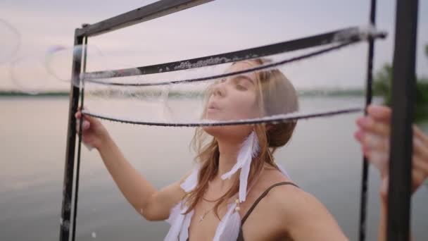 Uma jovem artista feminina sopra um monte de bolhas de sabão, mostra um show teatral usando uma moldura em câmera lenta ao pôr do sol em um lago — Vídeo de Stock