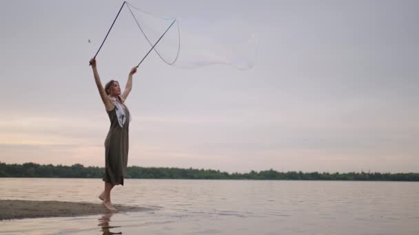 一个身穿裙子头戴羽毛的年轻嬉皮士女人在日落时分缓慢地在湖岸上制造巨大的肥皂泡 — 图库视频影像