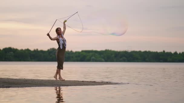 Una joven artista muestra un espectáculo de burbujas de jabón haciendo estallar enormes burbujas de jabón en la orilla de un lago al atardecer. Mostrar un hermoso espectáculo de burbujas de jabón en cámara lenta — Vídeo de stock