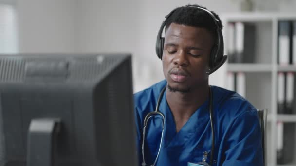 Un hombre negro se sienta en una computadora con un uniforme de médico y escribe una tarjeta de paciente mientras recibe llamadas con auriculares. Línea directa de ambulancia recibe llamadas y distribuye ambulancias — Vídeo de stock