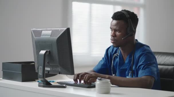 Czarny mężczyzna siedzi przy komputerze w mundurze lekarza i pisze kartkę pacjenta, odbierając telefony ze słuchawkami. Pogotowie ratunkowe Infolinia odbiera połączenia i rozsyła karetki — Wideo stockowe