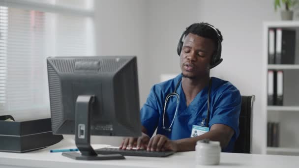 黒い男は医師の制服を着たコンピュータに座って、ヘッドフォンで電話をかけながら患者カードを書きます。救急車ホットラインは救急車を受け取り、配布します — ストック動画