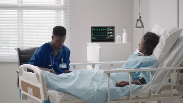 Egy férfi orvos beszél egy beteggel, aki egy kórházi ágyon fekszik egy kórházi kórteremben. Oxigénhez és maszkhoz csatlakoztatva. — Stock videók