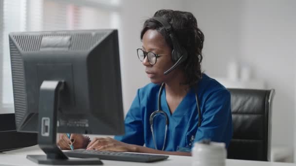 Лікар чорної жінки носить біле пальто, окуляри та гарнітуру, розмовляючи з клієнтом, використовуючи відеоколекцію. Через пандемічний спалах коронавірусу терапевт, який працює дистанційно, надає допомогу пацієнтам клініки за допомогою відеозв'язку — стокове відео