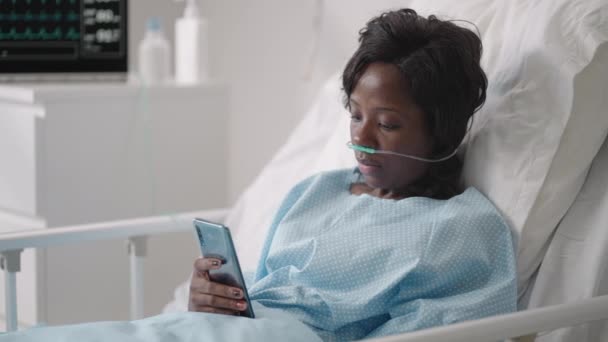 一位年轻妇女躺在医院病房时，在手机上留言。一名非洲女孩戴着面具躺在一个与心电图和氧气装置相连的病房里，给亲戚们留言 — 图库视频影像