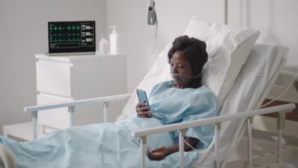 Een jonge vrouw schrijft een bericht op haar telefoon terwijl ze op een ziekenhuisafdeling ligt. Een Afrikaans meisje ligt op een afdeling aangesloten op ECG en zuurstof apparaten in een masker en schrijft berichten naar familieleden — Stockvideo