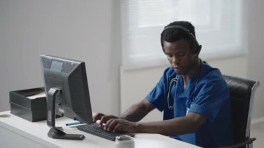 Siyahi bir adam doktor üniforması içinde bilgisayarın başında oturuyor ve kulaklıkla telefonlara bakarken hasta kartı yazıyor. Ambulans yardım hattı çağrıları alıyor ve ambulans dağıtıyor.