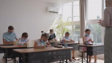 Sınıfta çalışan, sınıfta çalışan, öğretmenlerin açıklamalarını dinleyen ve yazan çok ırklı maskeler takan bir grup çocuk.