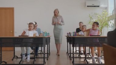 İlköğretim Okulu Sınıfı: Coşkulu Öğretmen Parlak Çeşitli Çocuklar Sıraları arasında yürüyor, Dersi Açıklıyor. Bir grup zeki çoklu etnik çocuk yeni şeyler öğreniyor. Yan Görünüm İzleme Görüntüsü