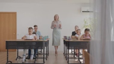 Sınıftaki çocuklar masalarında oturuyorlar ve öğretmen sınıfın etrafında yürüyor ve dersin konusunu açıklıyor. Akıllı Çocuklar Arkadaş Canlısı Modern Çevrede Öğreniyor