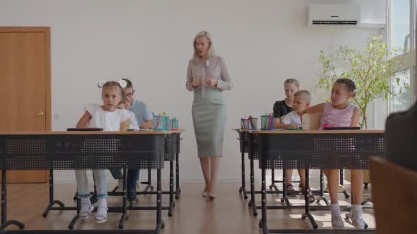 In de basisschoolklas: Enthousiaste leraar loopt tussen rijen van Bright diverse kinderen, verklaart les. Een groep slimme multi-etnische kinderen die nieuwe dingen leren. Side View Tracking Shot — Stockvideo