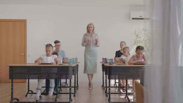 As crianças da turma estão sentadas em suas mesas e o professor caminha pela turma e explica o tema da aula. Crianças inteligentes aprendendo em ambiente moderno amigável — Vídeo de Stock