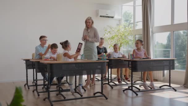 In Elementary School Class: Entusiasta insegnante cammina tra righe di luminoso Diversi bambini, Spiega Lezione. Gruppo di ragazzi intelligenti multietnici che imparano cose nuove. Vista laterale Tracking Shot — Video Stock