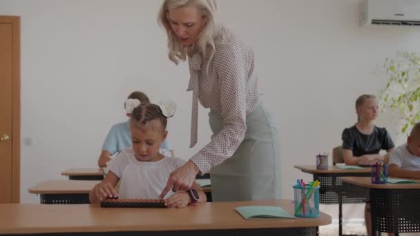 Die Kinder in der Klasse sitzen an ihren Schreibtischen und die Lehrerin geht durch die Klasse und erklärt das Unterrichtsthema. Intelligente Kinder lernen in freundlicher moderner Umgebung — Stockvideo