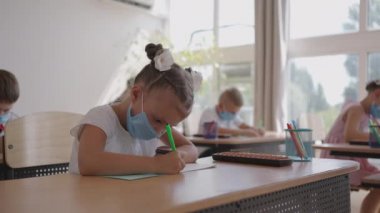 Corona virüsü salgını sırasında koruyucu maskeli bir öğrenci okulda ders çalışıyor. Maskeli bir öğretmen maskeli öğrencilere yeni bir ders konusunu açıklıyor. Çocuklar sınıfta kalemle yazarlar.