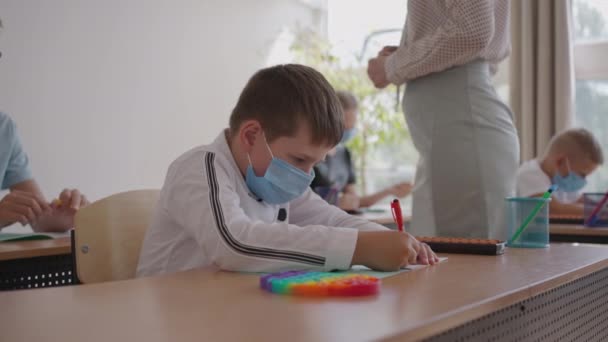 Grupo multirracial de niños con máscaras faciales que trabajan en clase, escribiendo y escuchando explicaciones del profesor en el aula — Vídeo de stock