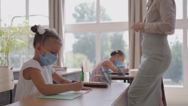Wielorasowa grupa dzieci w maskach pracujących w klasie, piszących i słuchających wyjaśnień nauczyciela w klasie — Wideo stockowe