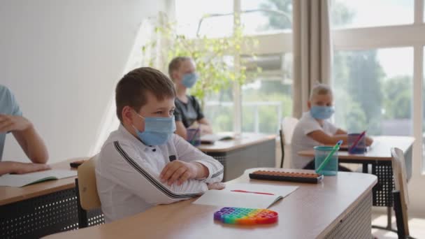 In der Schule sitzen maskierte Kinder im Klassenzimmer und beantworten die Fragen der Lehrer, indem sie in Zeitlupe die Hände heben. Unterricht während der Pandemie in der Schule — Stockvideo