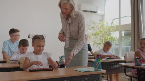 Lehrerin steuert Lernprozess in der Grundschule. Eine Lehrerin geht zwischen den Schreibtischen hin und her und kümmert sich um die Bearbeitung der Aufgabe — Stockvideo