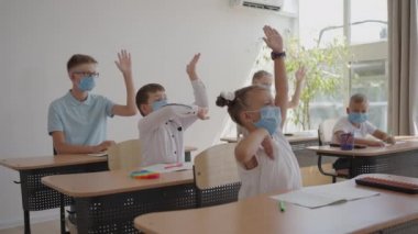 Sınıftaki maskeli çocuklar sınıfta oturur ve öğretmenlerin sorularını yavaş çekimde cevaplarlar. Okulda salgın sırasında alınan dersler