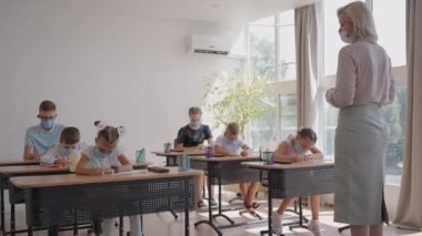 Corona virüsü salgını sırasında koruyucu maskeli bir öğrenci okulda ders çalışıyor. Maskeli bir öğretmen maskeli öğrencilere yeni bir ders konusunu açıklıyor. Çocuklar sınıfta kalemle yazarlar.