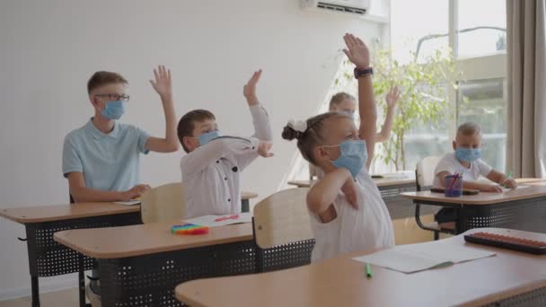 Діти в класі в школі в масках сидять в класі і відповідають на питання вчителів, піднімаючи руки в повільному русі. Уроки під час пандемії в школі — стокове відео